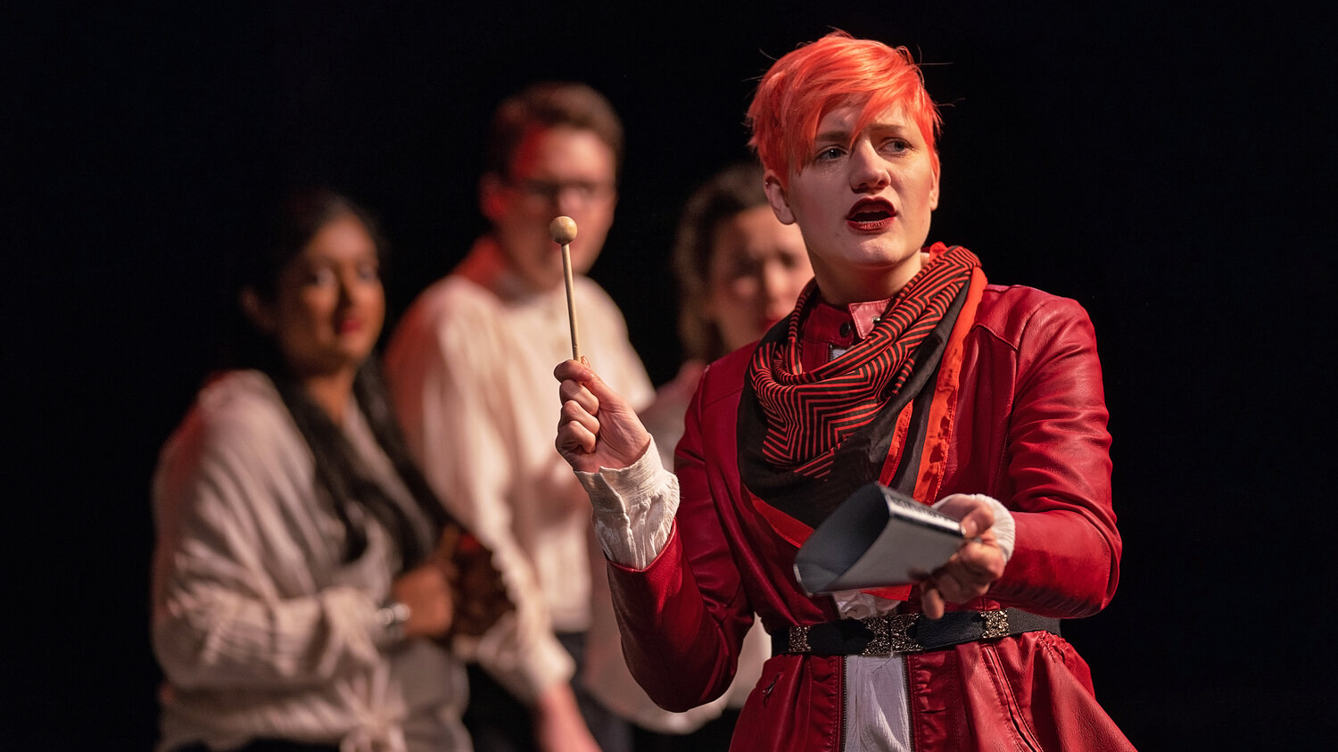 Actors perform in Romeo and Juliet. One actor faces the camera striking a cowbell while three others look on.