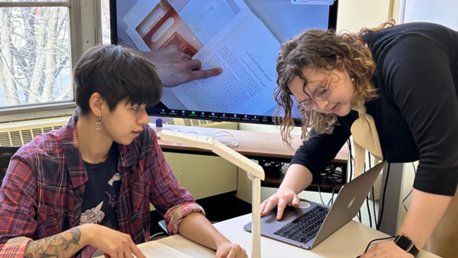Two students point at an open book, whose image is projected onto a large screen behind them