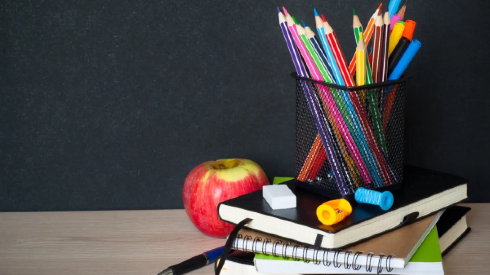 Stack of notebooks with a cup of colored pencils on top of it and an apple beside it. 