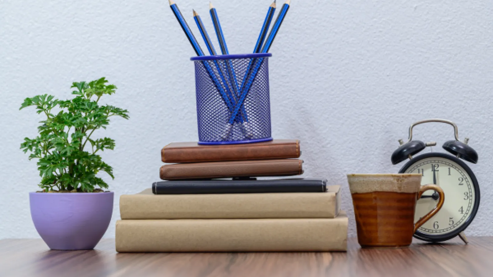 A stack of books with a pencil cup on top of them. To the left is a plant and to the right is a mug and clock.