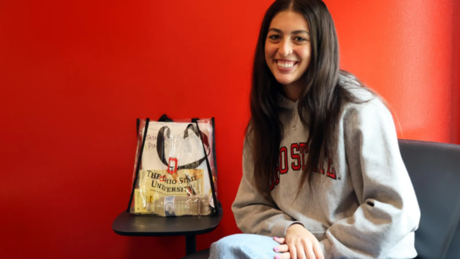 Jasmyn Maingi sits in front of a bright scarlet wall and smiles at the camera