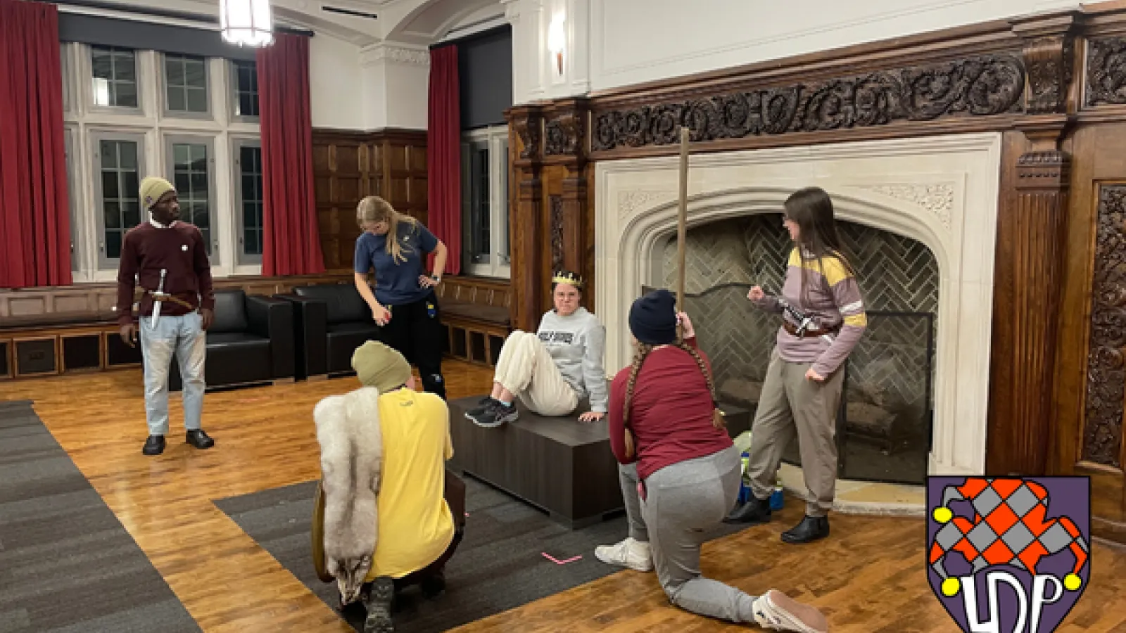 Six actors rehearse in front of fireplace. Some carry daggers, and one wears a crown.