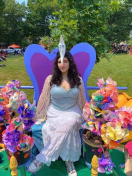 Celeste Alva Jimenez wearing her moon head piece seated outside on a colorful chair among rainbow paper flowers at Parade the Circle