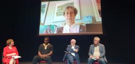 Four presenters sit onstage with one presenter projected on a screen behind them