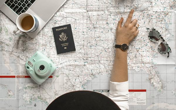 Camera, passport and sunglasses on top of a map with a person pointing