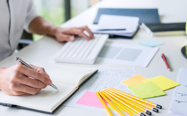 A hand writing in notebook on a desk full of office supplies