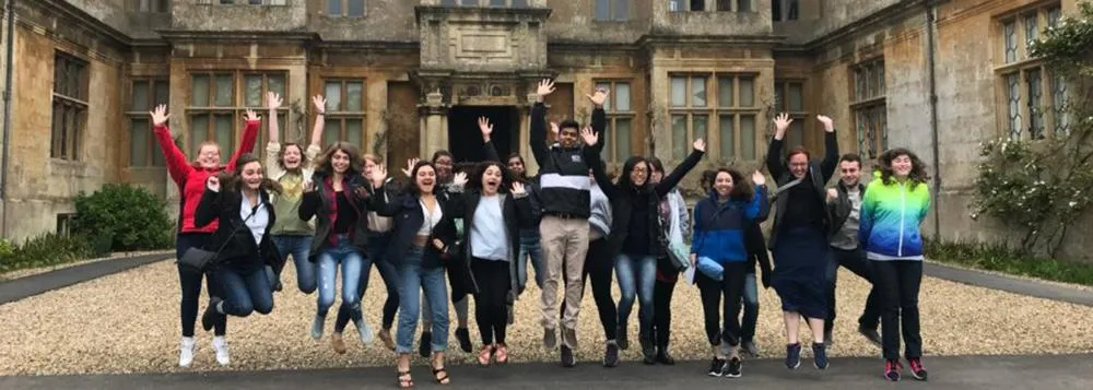 A group of students jumps and cheers together outside a historic stone building.