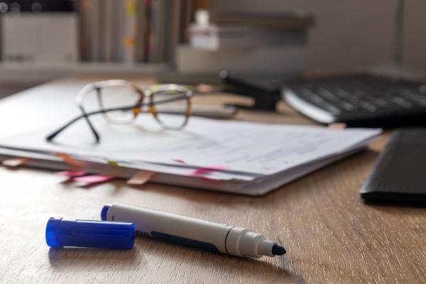 A blue marker, uncapped, sits on a wooden desk, with a pile of papers, a pair of glasses, a keyboard, and a stack of books in the background