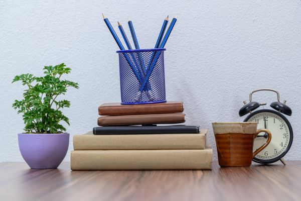 A stack of books with a pencil cup on top of them. To the left is a plant and to the right is a mug and clock.