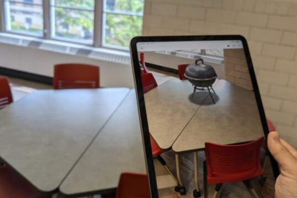 A handing holding up an iPad whose screen shows a VR grill sitting on the table behind the iPad.