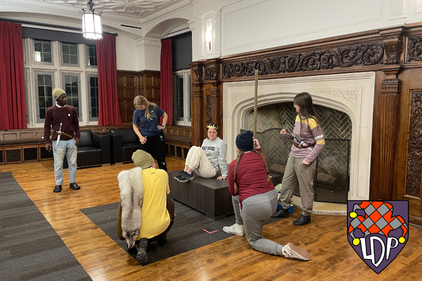 Six actors rehearse in front of fireplace. Some carry daggers, and one wears a crown.