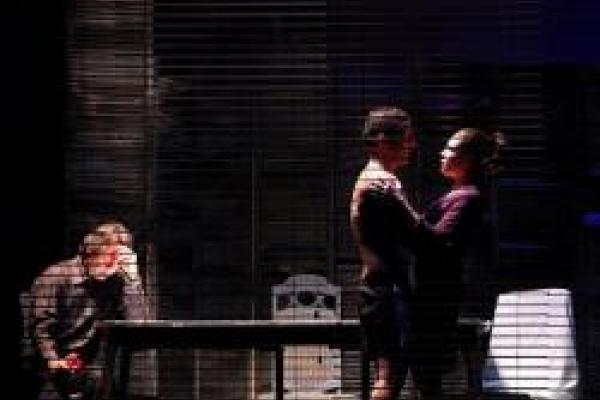 On a theatre stage, a couple embraces while a third person sits at a table with their head resting on their hand