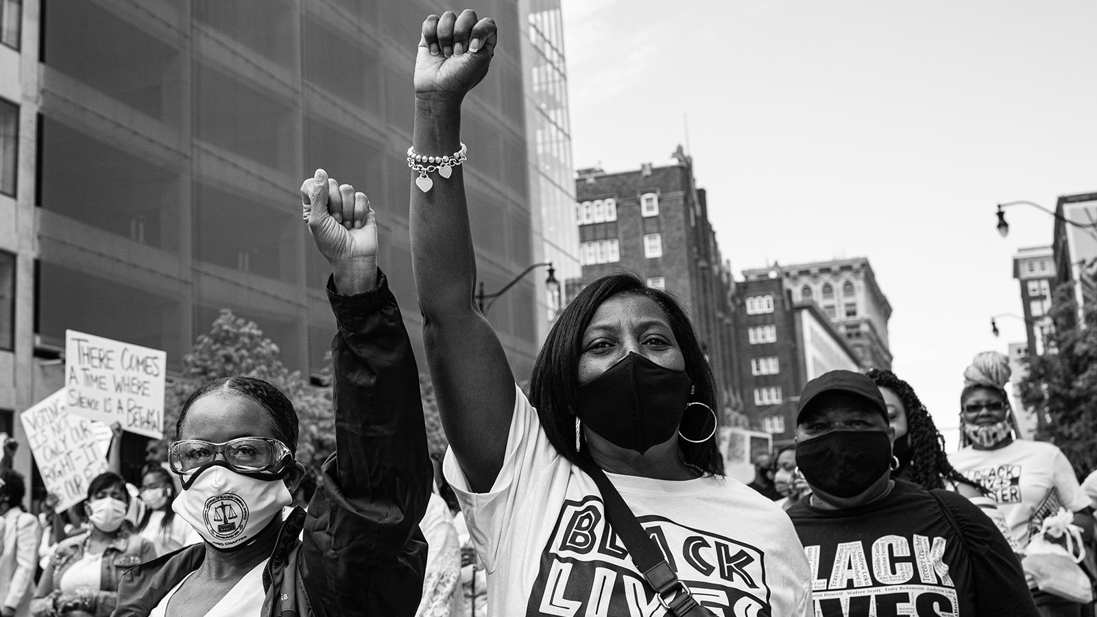 Three Black protestors stand in a street, masked. Two have arms raised