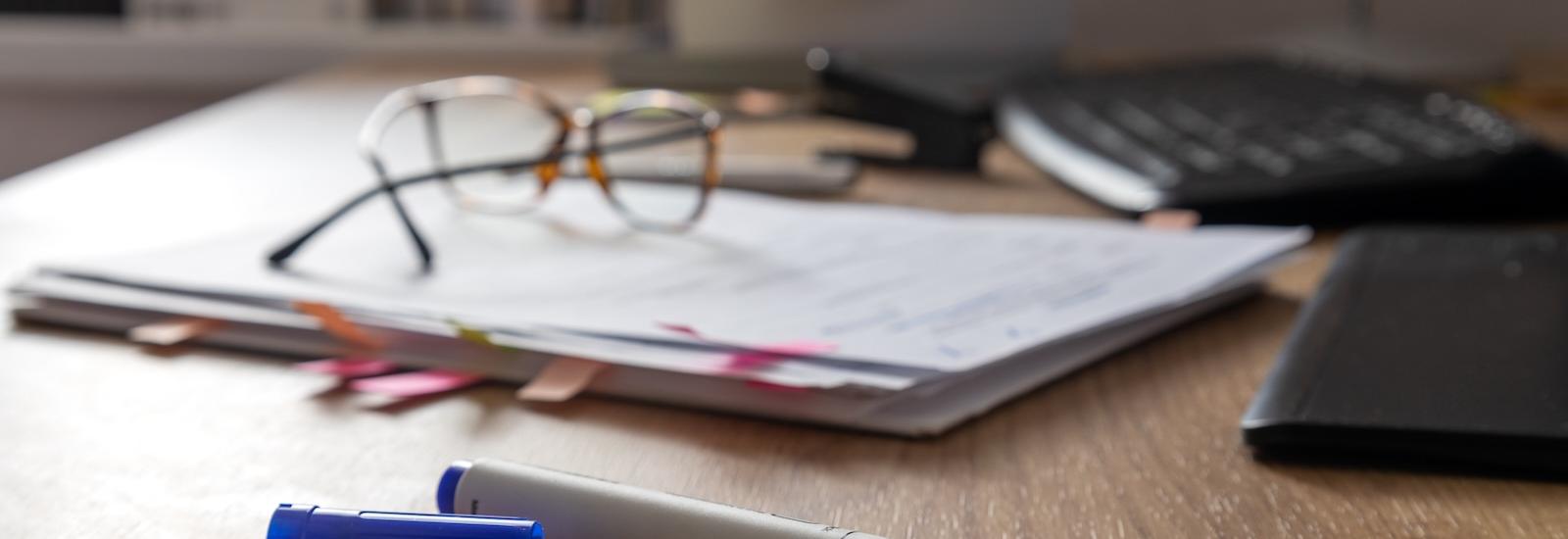 A blue marker, uncapped, sits on a wooden desk, with a pile of papers, a pair of glasses, a keyboard, and a stack of books in the background